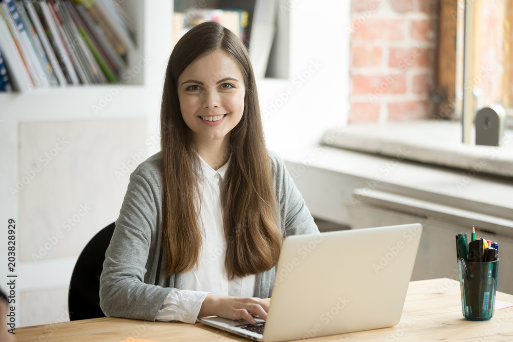 Portrait of smiling Caucasian female worker looking to camera while working at laptop in modern office. Student posing for academic class book. Concept of confidence, success, achievements Portrait of smiling Caucasian female worker looking to camera while working at laptop in modern office. Student posing for academic class book. Concept of confidence, success, achievements