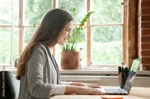 Happy female office associate smiling while working at laptop, reading good business news of company success. College student satisfied with email notification about high exam mark.