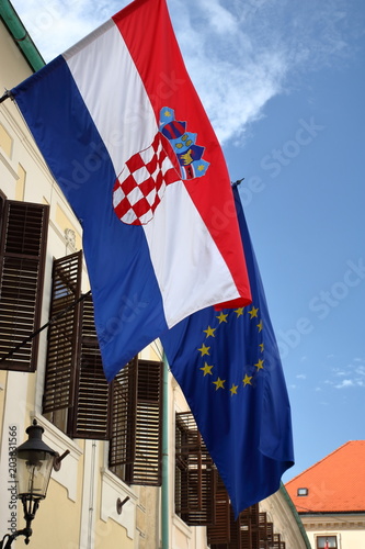 Flaga Chorwacja wisi na ścianie budynku, na zewnątrz, z nią wisi flaga Unii europejskiej, w budynku otwarte okiennice, błękitne niebo w tle