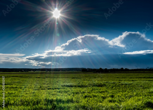 bright sun shines over clouds and flat green field, forest long away on horizon line