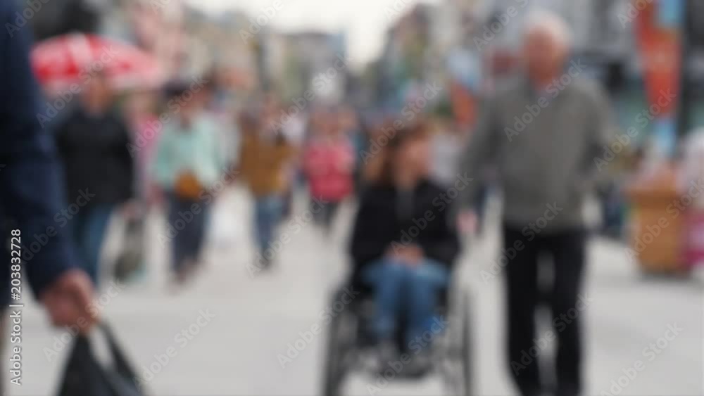 Anonymous disabled people in wheelchair on street. Blurred lady in wheelchair being pushed along crowded city street. Out of focus background from city with people in weekend Anonymous disabled people in wheelchair on street. Blurred lady in wheelchair being pushed along crowded city street. Out of focus background from city with people in weekend