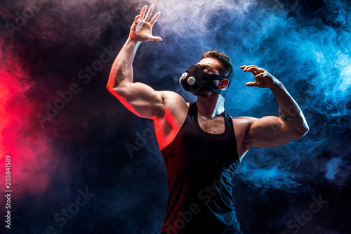 Strong male athlete in a black training mask posing on a black background with smoke photo
