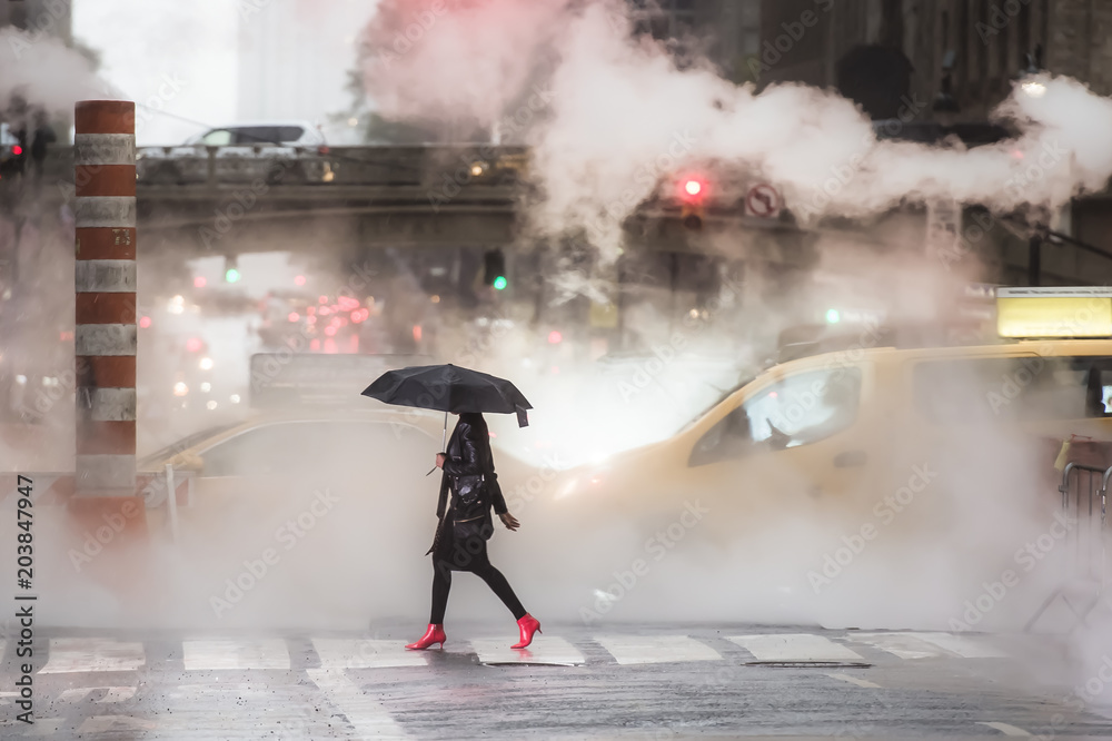 A woman with an umbrella and red high heels shoes is crossing the 42nd street in Manhattan. Taxi and steam coming out from from the manholes in the background. New York City, Usa.