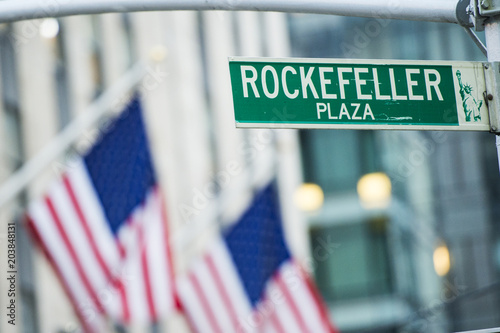 Close-up view of green street sign depicting it is Rockefeller Plaza in Midtown Manhattan, New-York. Blurred American flags in the background