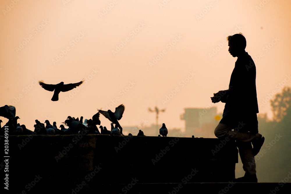 Silhouette of an Indian boy who is feeding some pigeons in a Ghat in Varanasi during a beautiful sunset, India
