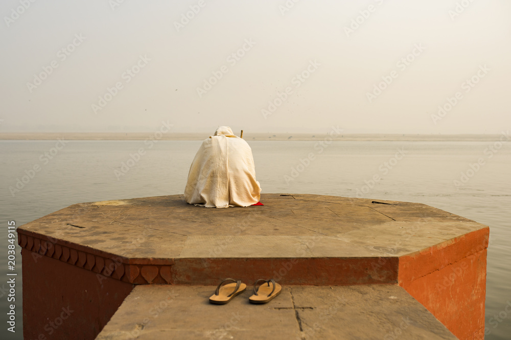 A holy man dressed in white is praying and meditating on one of the many Ghats of Varanasi in front of the sacred river Ganges, India.