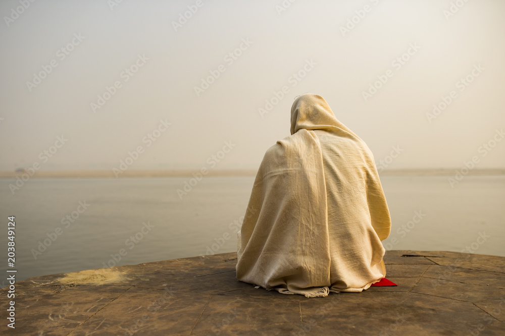 A holy man dressed in white is praying and meditating on one of the many Ghats of Varanasi in front of the sacred river Ganges, India.