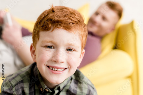 close-up view of cute redhead boy smiling at camera while spending time with father at home