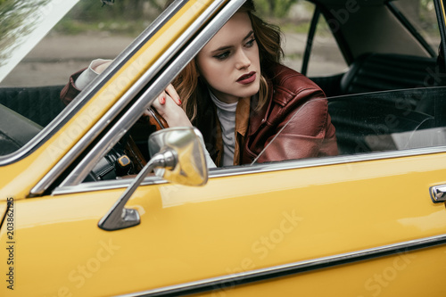 attractive girl leaning at steering wheel and looking away while sitting in yellow vintage car