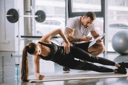 Male personal trainer looking at timer and young athletic woman doing side plank on fitness mat