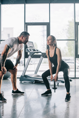 male personal trainer looking at sportswoman doing exercises with dumbbell at gym