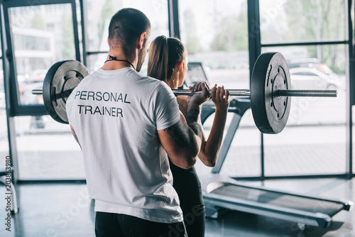 male personal trainer helping sportswoman to do exercises with barbell at gym