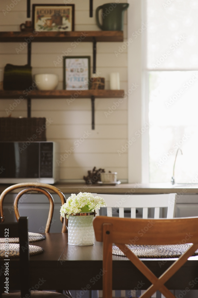 cozy summer morning at rustic country house kitchen. Wooden table with bouquet of fresh flowers, open shelving. Casual breakfast in grey and brown interior.