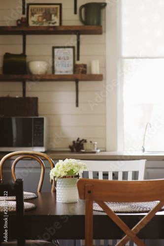 cozy summer morning at rustic country house kitchen. Wooden table with bouquet of fresh flowers  open shelving. Casual breakfast in grey and brown interior.