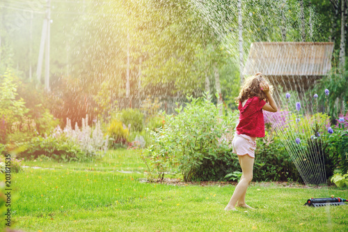 Kid girl playing with garden sprinkler in hot summer day. Water outdoor activities for children, happy childhood.