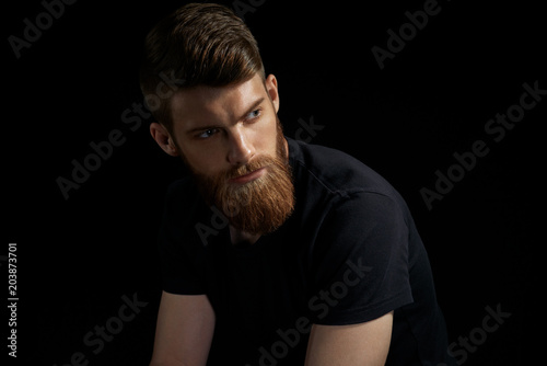 Portrait of handsome single bearded young man with serious expression and black shirt looking over black background with copy space