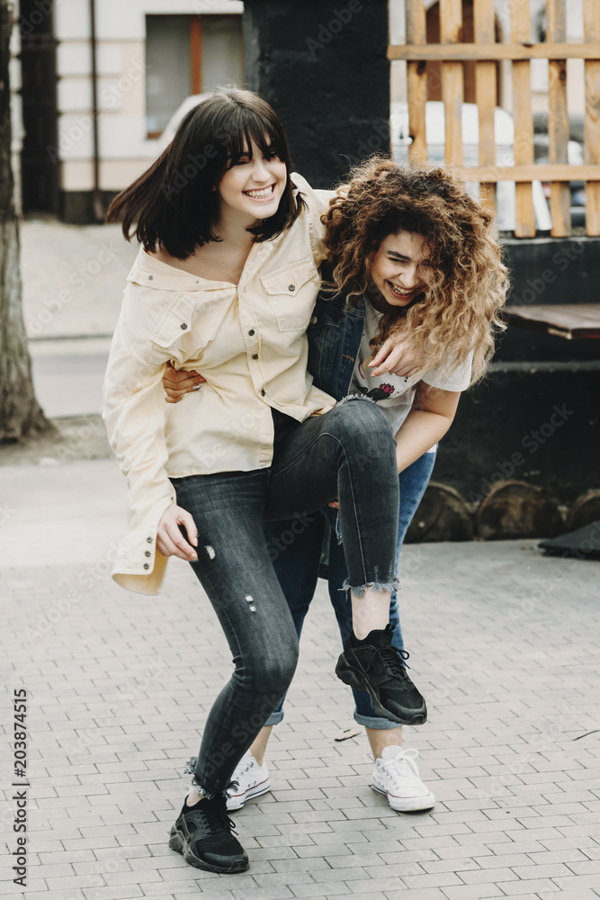 Full length portrait of two funny females laughing where curly girl is trying to hold on her arms her girlfriend brunette.