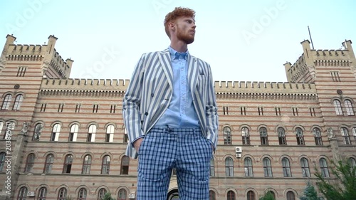 Confident attractive man with curly ginger hair in trews pants and striped jacket walks at the background of the old building. He stops, looks away and goes back. photo
