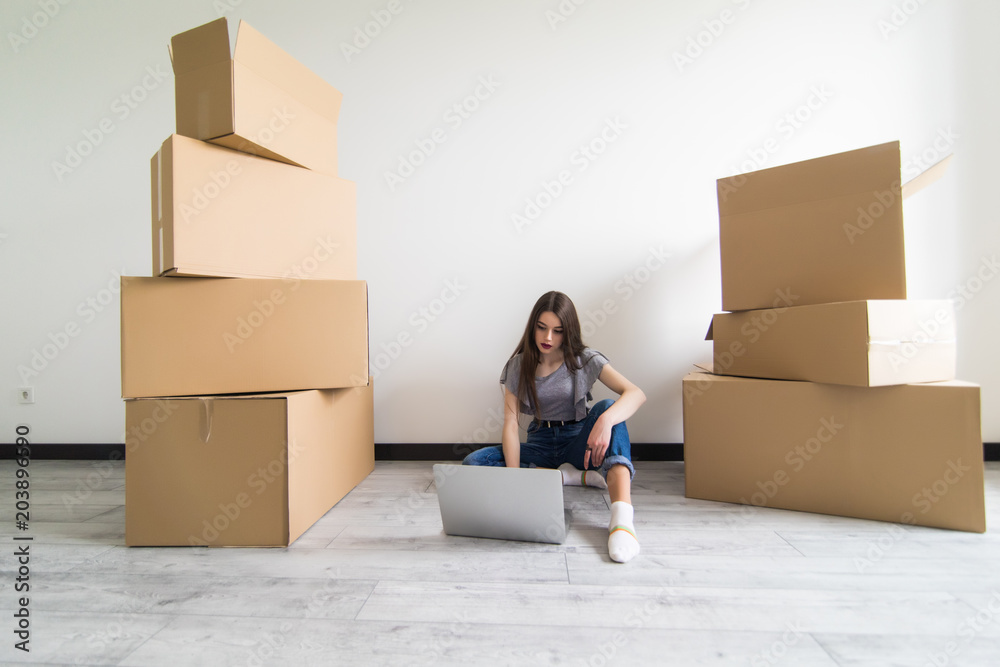 Happy young woman sitting on floor near moving boxes. Young woman moving to new home. Woman using laptop, looking at camera and smiling Happy young woman sitting on floor near moving boxes. Young woman moving to new home. Woman using laptop, looking at camera and smiling