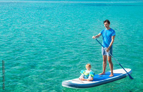Young Father with His Son on SUP. Dad and Son Concept. Man and Boy Stand Up Paddling. Blue Sea Water. Floating Sup Board. Dad and Son on Summer Vacation Concept. Copy Space.