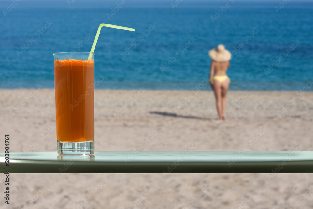 A glass of carrot juice with a straw in the cafe. Female in the bikini and straw hat on the beach
A glass of carrot juice with a straw in the cafe. Female in the bikini and straw hat on the beach