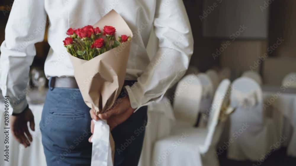 Loving man is hiding red roses behind his back bringing beautiful bouquet for his date in restaurant. Flowers, romantic relationship and dating concept. Loving man is hiding red roses behind his back bringing beautiful bouquet for his date in restaurant. Flowers, romantic relationship and dating concept.