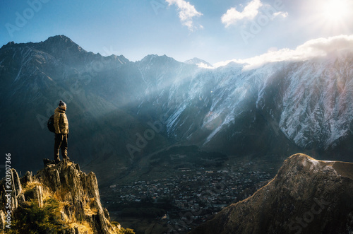 Young tourist in bright hat, black trousers with a backpack stands on cliff's edge and looking at the misty mountain village and glacier at sunrise, Stepantsminda, Georgia