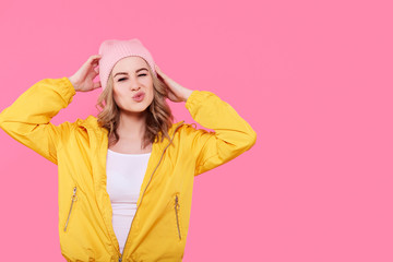 Crazy beautiful trendy girl in bright yellow jacket and pink beanie hat puckering lips. Attractive cool young woman fashion portrait over pastel pink background.