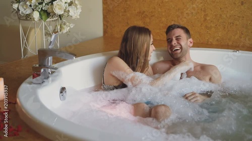 Pretty cheerful girl is blowing soap foam and having fun with her bearded fience in hot tub in day spa. Young people are laughing, enjoying weekend and talking. photo