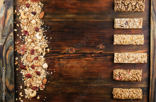 Variety of granola energy bars in row with scattered mixed nuts, cereals, cranberry on brown wood textured table background. Healthy nutritious vegan fitness food snack. Top view, copy space, close up