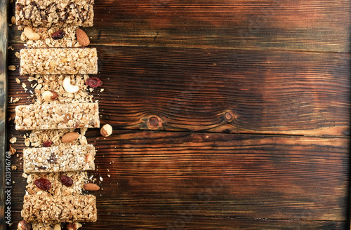 Variety of granola energy bars in row with scattered mixed nuts  cereals  cranberry on brown wood textured table background. Healthy nutritious vegan fitness food snack. Top view  copy space  close up