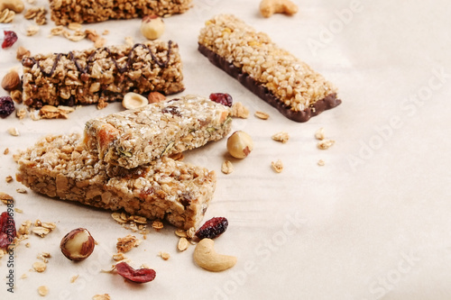Various granola chocolate energy bars in row with scattered mixed nuts, cereals & dried fruit, grunged white wood table background. Healthy vegan fitness food snack. Top view, copy space, close up.