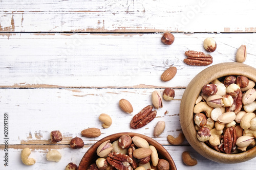 Mixed nuts in wooden bowl and scattered on table. Trail mix of pecan, almond, macadamia & brazil edible nuts with walnut hazelnut on wood textured surface. Background, copy space, top view, close up.