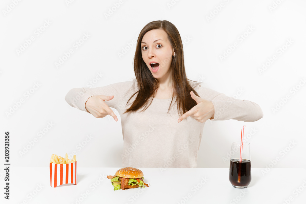 Beautiful young woman sitting at table with burger, french fries, cola in glass bottle isolated on white background. Proper nutrition or American classic fast food. Advertising area with copy space. Beautiful young woman sitting at table with burger, french fries, cola in glass bottle isolated on white background. Proper nutrition or American classic fast food. Advertising area with copy space.