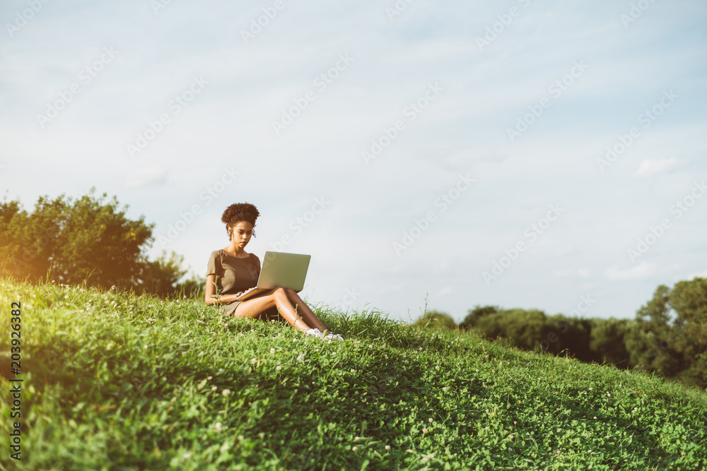 A pensive young African-American woman is sitting alone on the grass of the flank of the hill and using her netbook on a sunny summer day; black girl with the netbook outdoors on the meadow A pensive young African-American woman is sitting alone on the grass of the flank of the hill and using her netbook on a sunny summer day; black girl with the netbook outdoors on the meadow