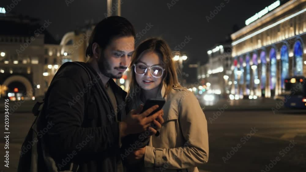 two young female male friends chatting and looking at their smartphone at street night, man and woman happy student tourists two young female male friends chatting and looking at their smartphone at street night, man and woman happy student tourists