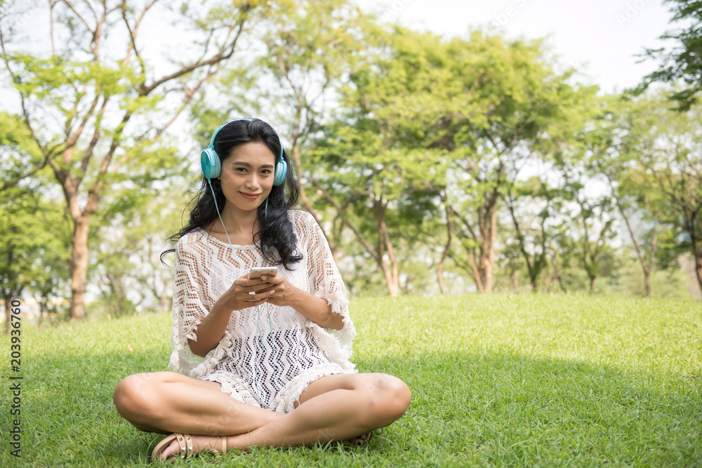 Portrait of beautiful Asian tan Bohemian girl with hipster style dress playing smartphone or mobile phone in park. Female listening music on garden field. Relax in weekend. Portrait of beautiful Asian tan Bohemian girl with hipster style dress playing smartphone or mobile phone in park. Female listening music on garden field. Relax in weekend.