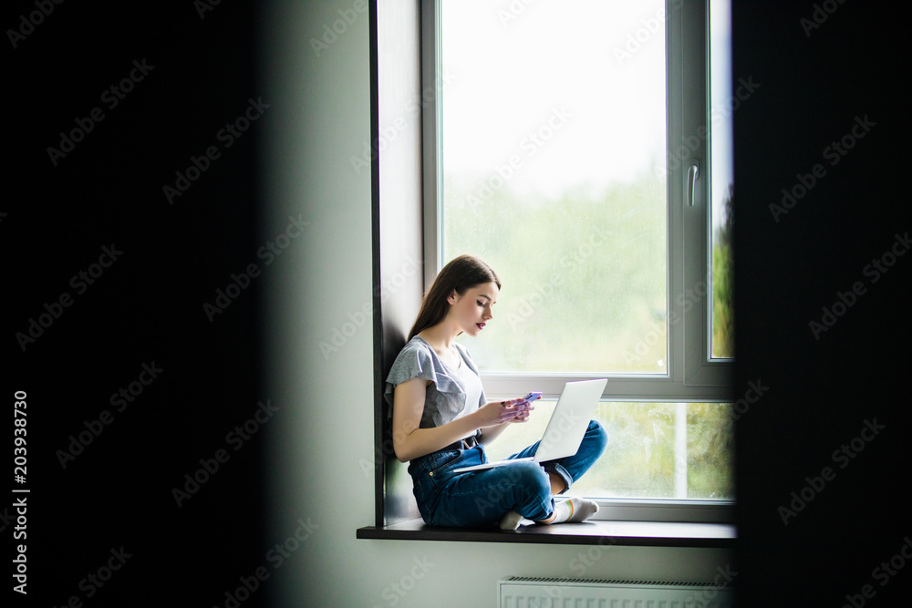 Beautiful young woman sitting on windowsill and using laptop and typing on phone