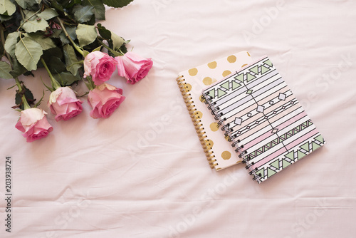 Floral frame with stunning pink roses and notebooks on pink bed sheets in the bedroom. Freelance workspace. Wedding, gift card, valentine's day or mother's day background