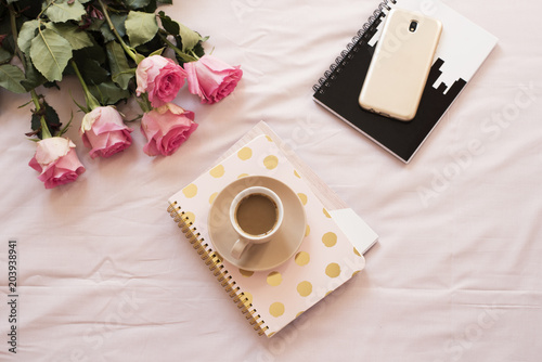 Coffee in bed on pink sheets. Roses, notebooks, gold smartphone around. Freelance fashion home femininity workspace
