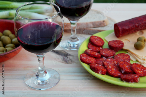 Food and beverages - still life - two glasses of red wine on a table with sliced salami, cheese, bread and green olives