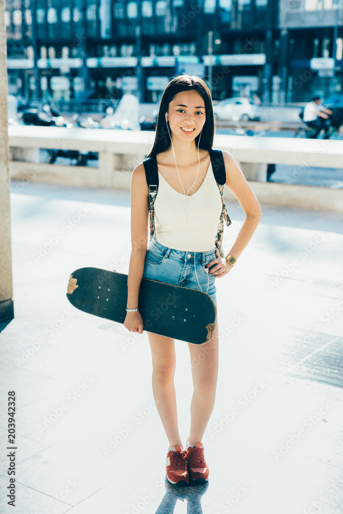 young woman posing outdoor back light holding skateboard looking camera smiling listening music - youth culture, skater, rebel concept