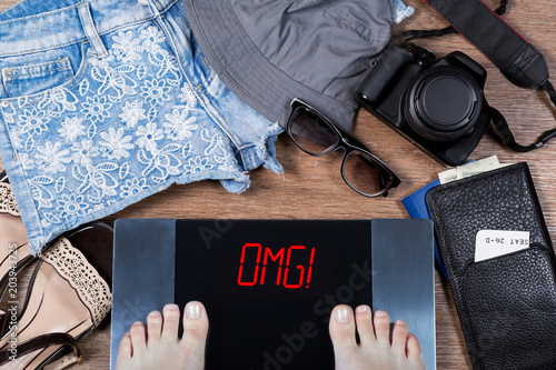 Woman weighs herself before vacation. Concept of unhealthy lifestyle and weight control. Digital scales with sign omg! and holiday accessories. Top view.