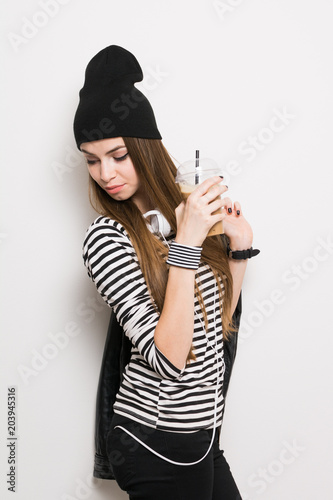 Teenage girl in black and white outfit with beanie, headphones, holding takeaway coffee. Studio lighting, no retouch, no filter.