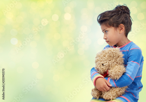 childhood, sadness, loneliness and people concept - sad little girl with teddy bear toy over green holidays lights background photo