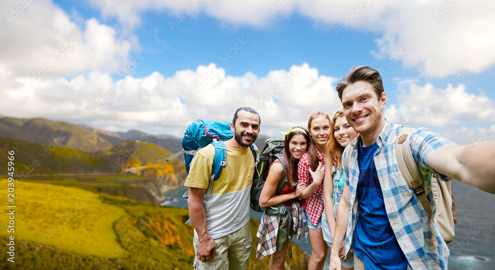 technology, travel, tourism, hike and people concept - group of smiling friends with backpacks taking selfie over big sur coast of california background