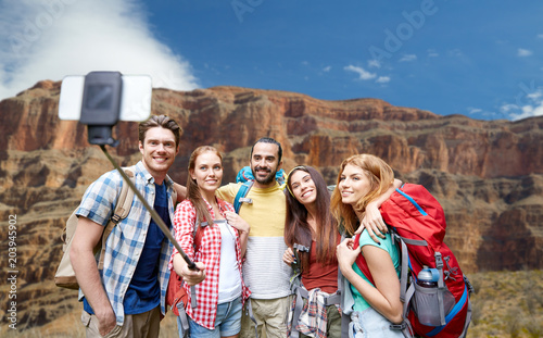 travel, tourism and technology concept - group of smiling friends walking with backpacks taking picture by smartphone on selfie stick over rocks of grand canyon national park background