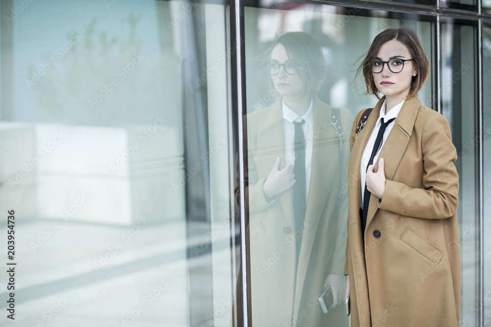 Manager con occhiali da vista vestita con cappotto beige , camicia bianca e cravatta nera , cammina lungo una parete di vetro in cui si riflette Manager con occhiali da vista vestita con cappotto beige , camicia bianca e cravatta nera , cammina lungo una parete di vetro in cui si riflette