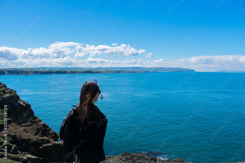 young woman with dark hair and backpack standing on cliff and looking on ocean, travelling concept
