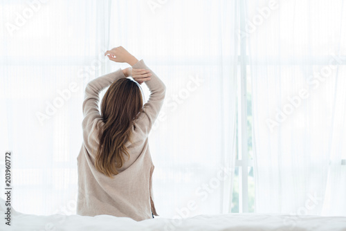 Wallpaper Mural Back view of happy beautiful young Asian woman waking up in morning, sitting on bed, stretching in cozy bedroom, looking through window. Funny woman after wake up. She is stretching and smiling. Torontodigital.ca